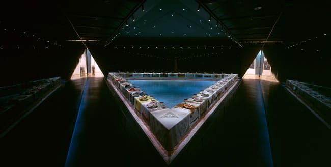 Triangular table with place settings and art pieces in dimly lit room, featuring a central blue area and patterned walls.