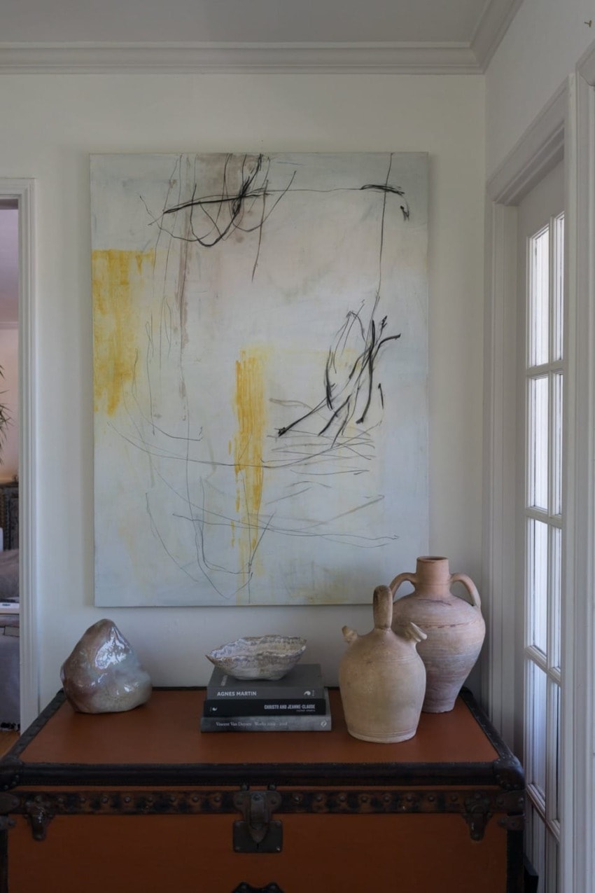 Abstract painting above a vintage trunk with pottery and books in a well-lit room.