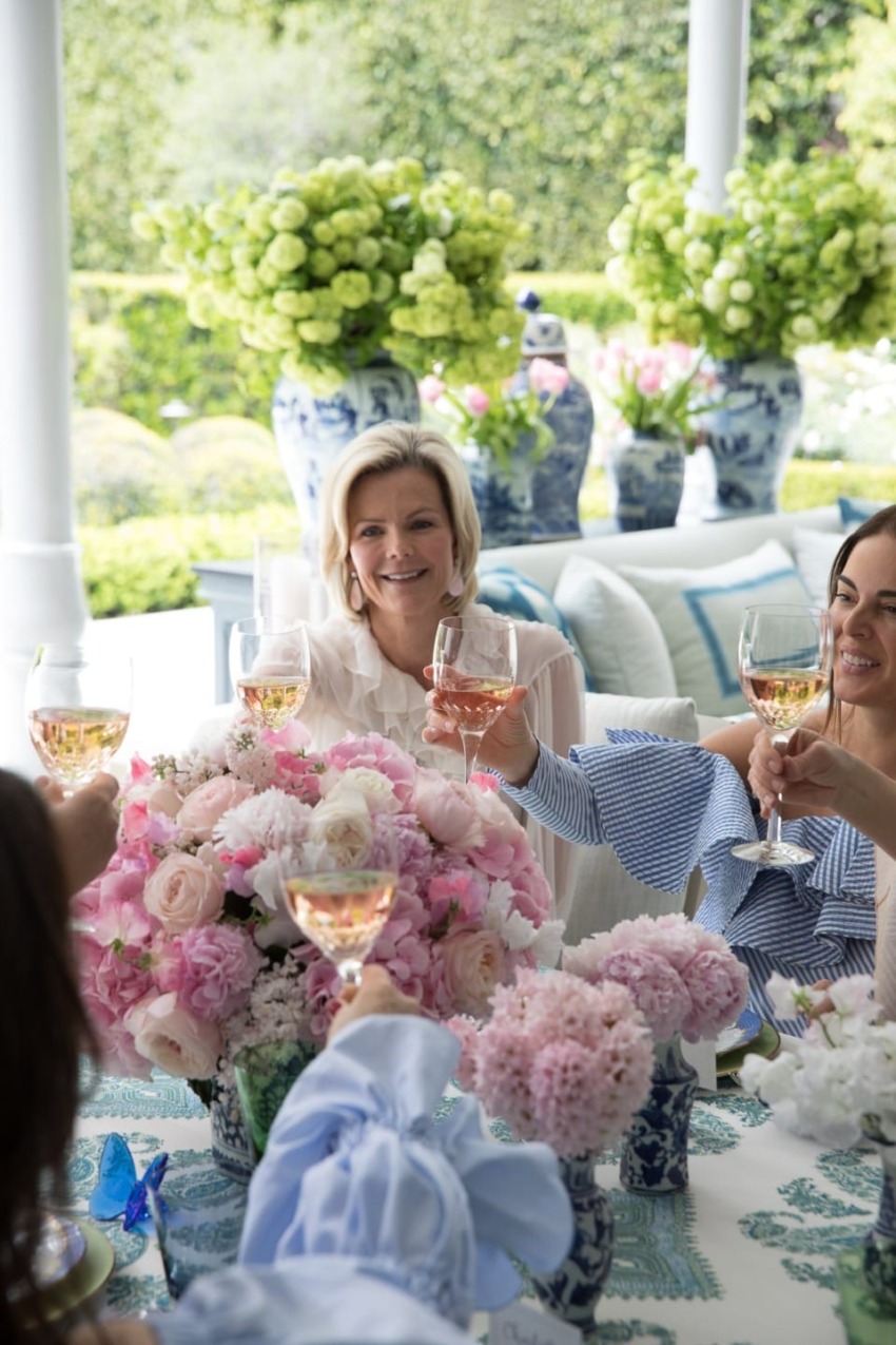 Women enjoying a toast with wine in a garden setting, surrounded by flowers and decorative vases.