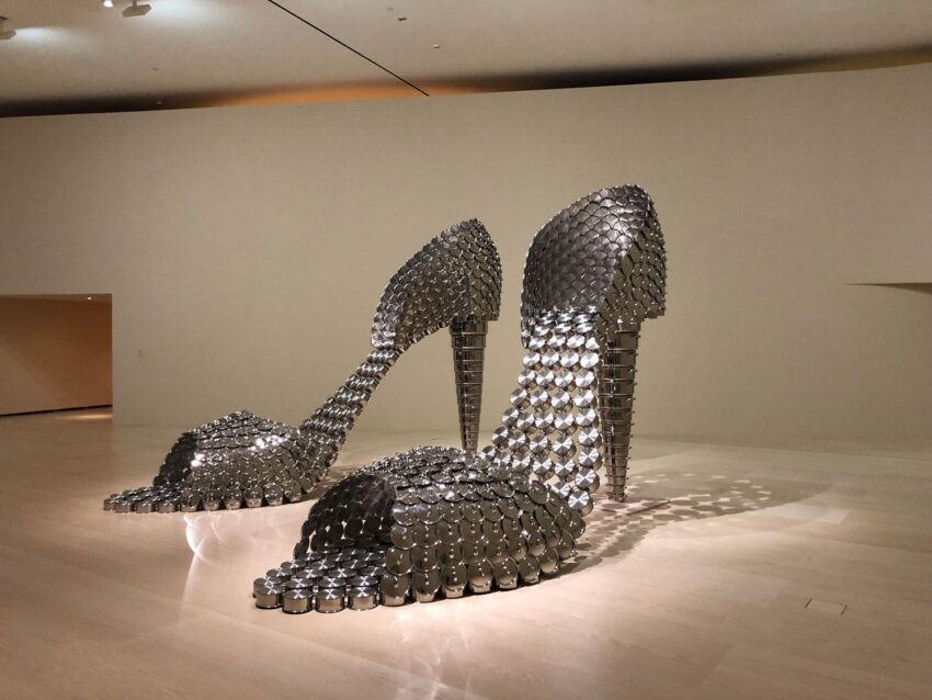 Sculpture of high-heeled shoes made from metal pots and lids displayed on a gallery floor.