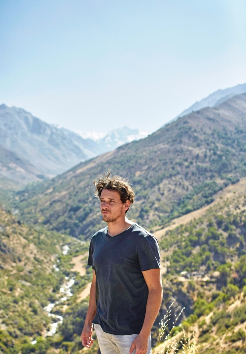 Man standing on a scenic mountain landscape with green valleys and distant snowy peaks under a clear blue sky.