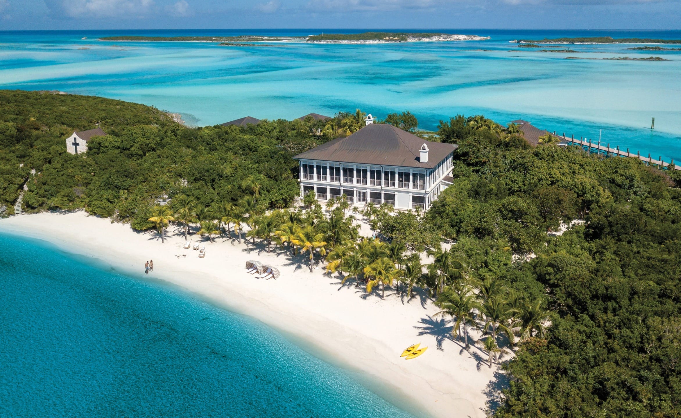 Aerial view of a beachfront house surrounded by trees, with a pristine sandy beach and turquoise ocean water.