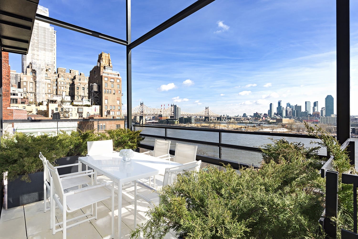 Rooftop terrace with a white dining set, overlooking a city skyline and bridge under a clear blue sky.