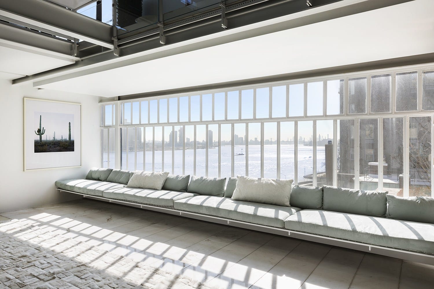 Spacious room with long couch, large windows, and cityscape view across a river on a bright day.