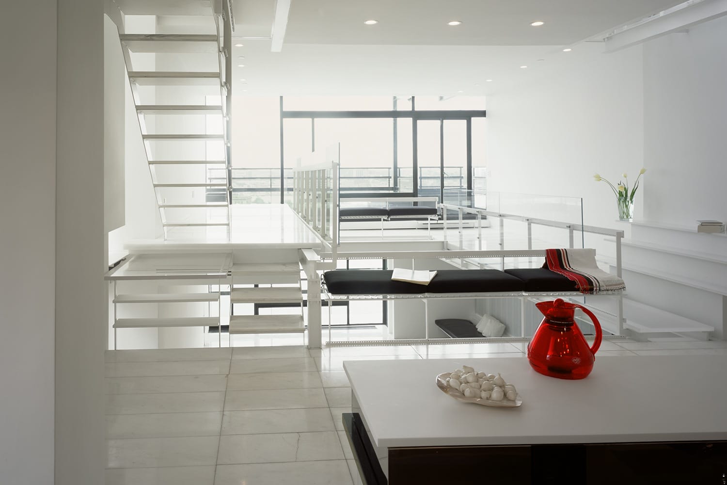 Modern loft interior with minimalist white decor, open staircase, and large windows. Red pitcher adds a pop of color.