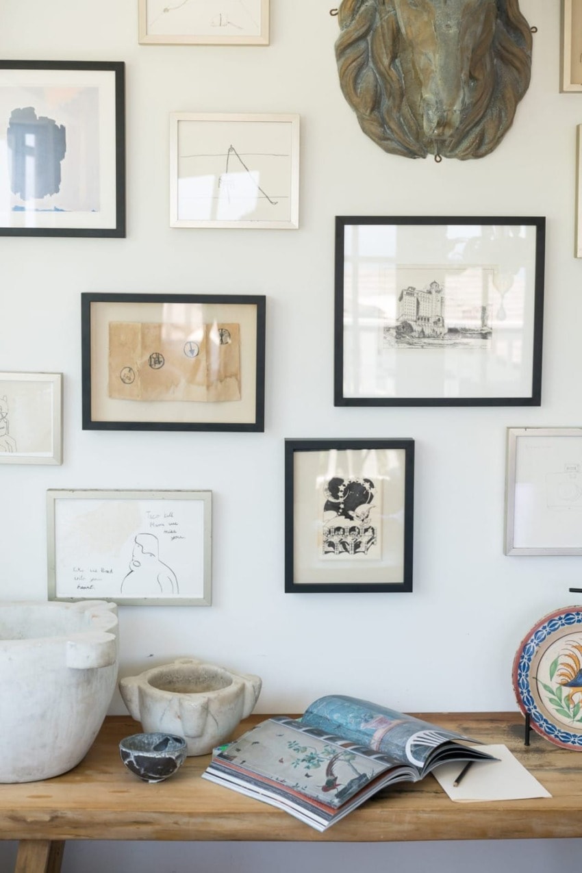 Gallery wall with various framed art, sketches, and a sculpture above a wooden table with open book and decorative bowl.