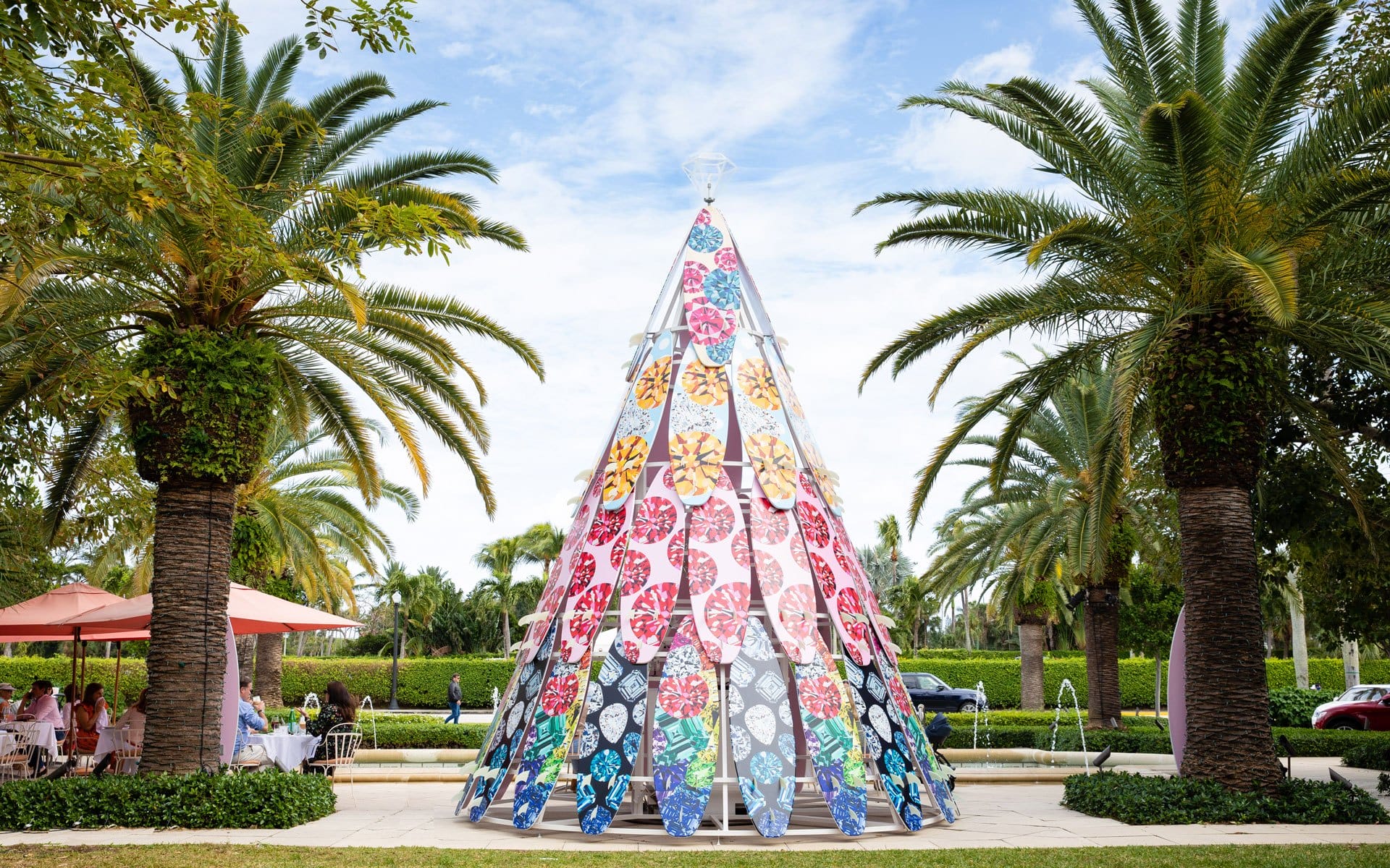 Colorful cone-shaped art installation surrounded by palm trees and outdoor dining area under a blue sky.