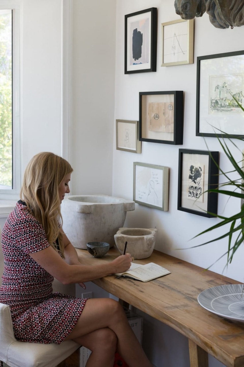 Woman writing in a journal at a wooden desk with framed art on a white wall in a bright room.