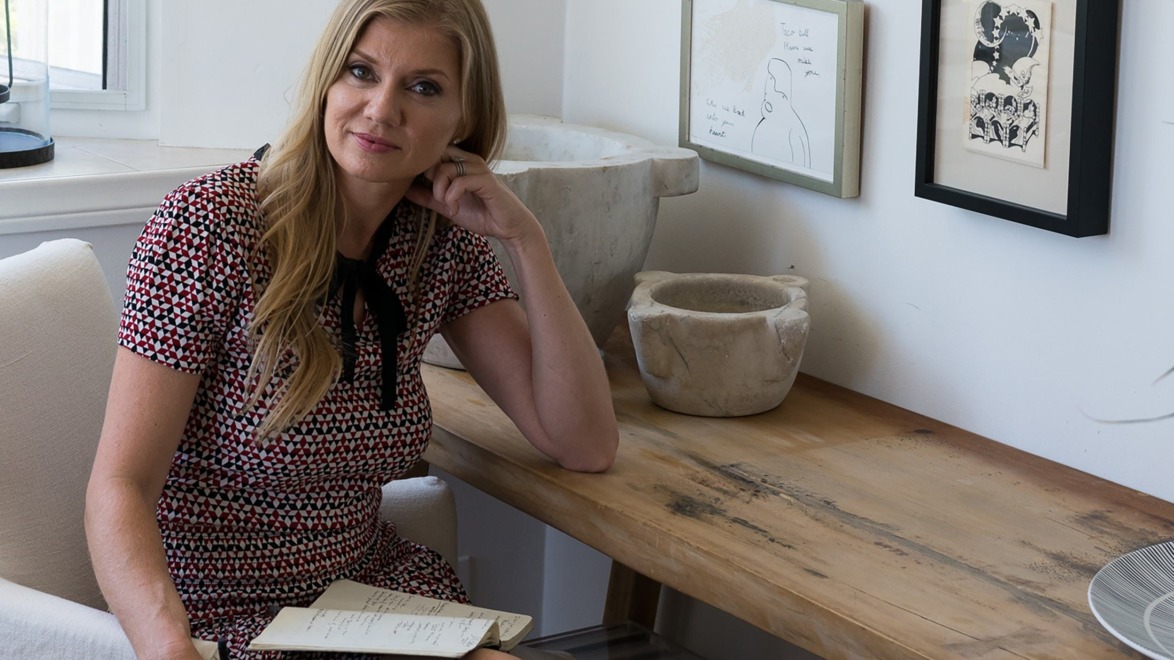 Woman sitting at a wooden desk with framed art on the wall, holding a notebook, wearing a patterned dress and smiling.