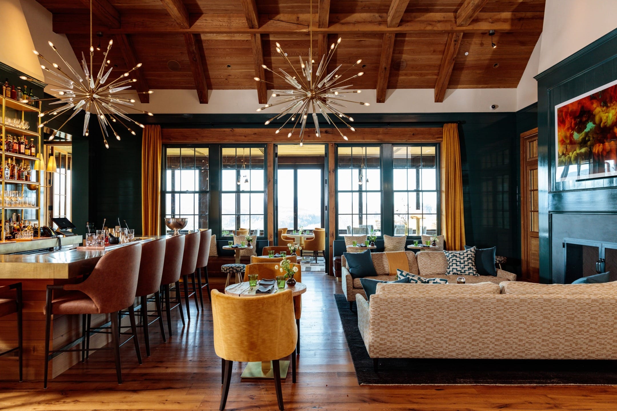 Elegant restaurant interior with wooden beams, plush seating, starburst chandeliers, and large windows offering scenic views.