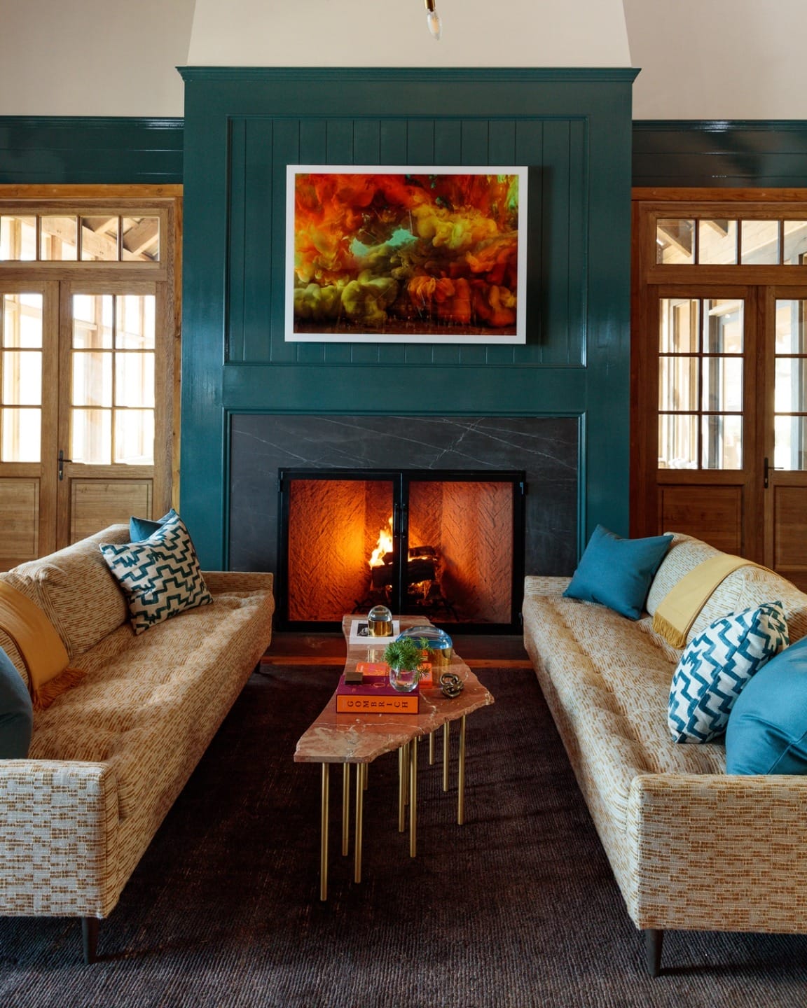 Cozy living room with a lit fireplace, two beige sofas, colorful cushions, and a vibrant painting above the mantel.