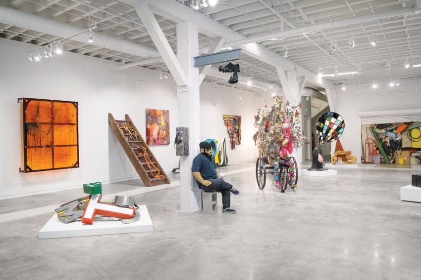 Art gallery with colorful sculptures and paintings displayed on walls and floor, featuring a person sitting on a bench.