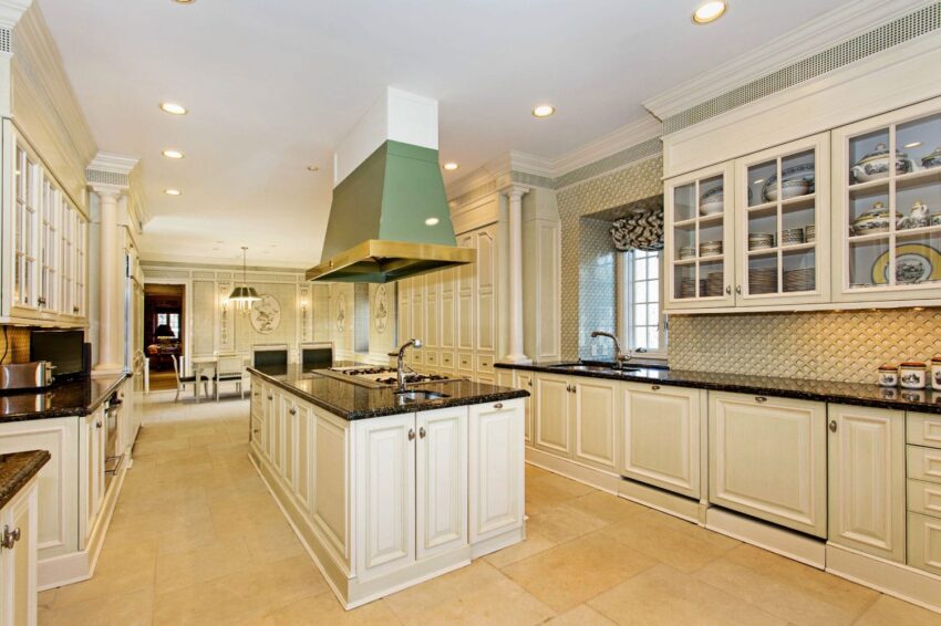 Spacious kitchen with cream cabinets, black countertops, green range hood, and tile backsplash under recessed lighting.