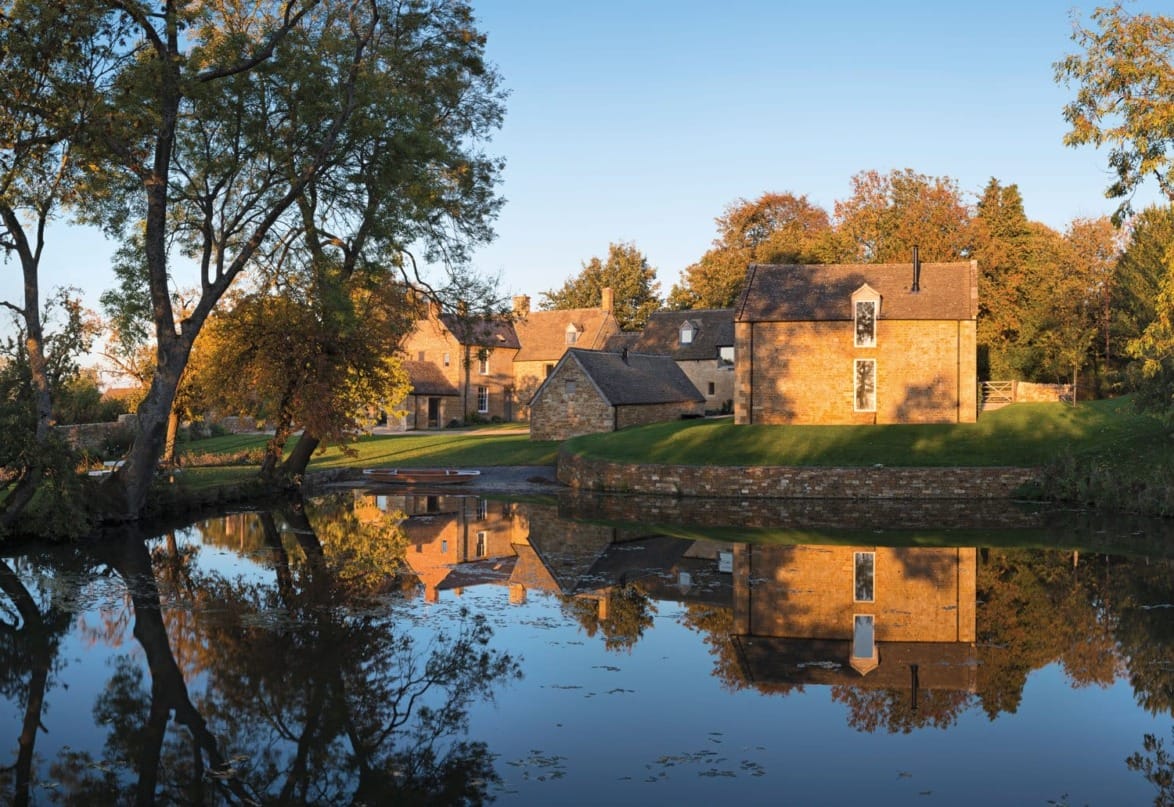 Scenic view of historic stone houses reflecting in a tranquil pond, surrounded by autumn trees under a clear blue sky.