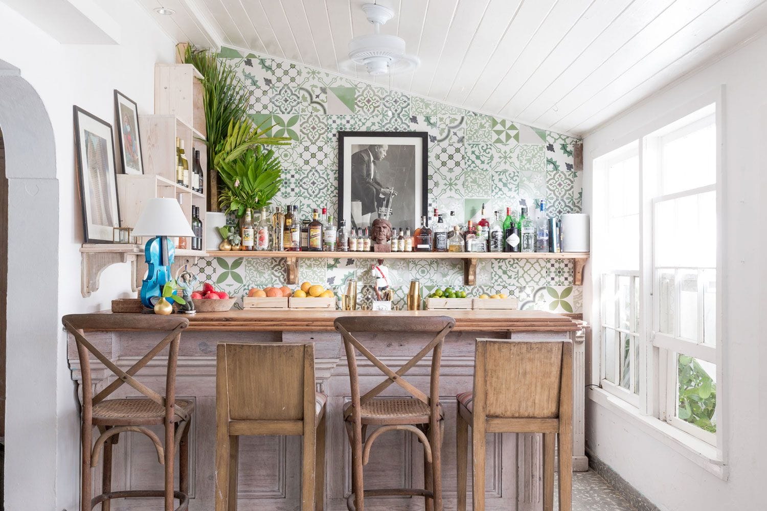 Rustic home bar with wooden stools, patterned tile backsplash, and shelves filled with bottles and plants. Bright natural light.