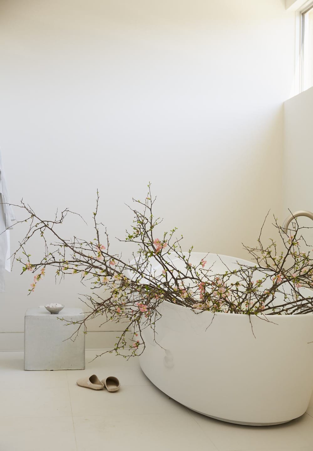 Minimalist bathroom with a white freestanding tub, decorative branches, slippers, and a small stool against a bright wall.