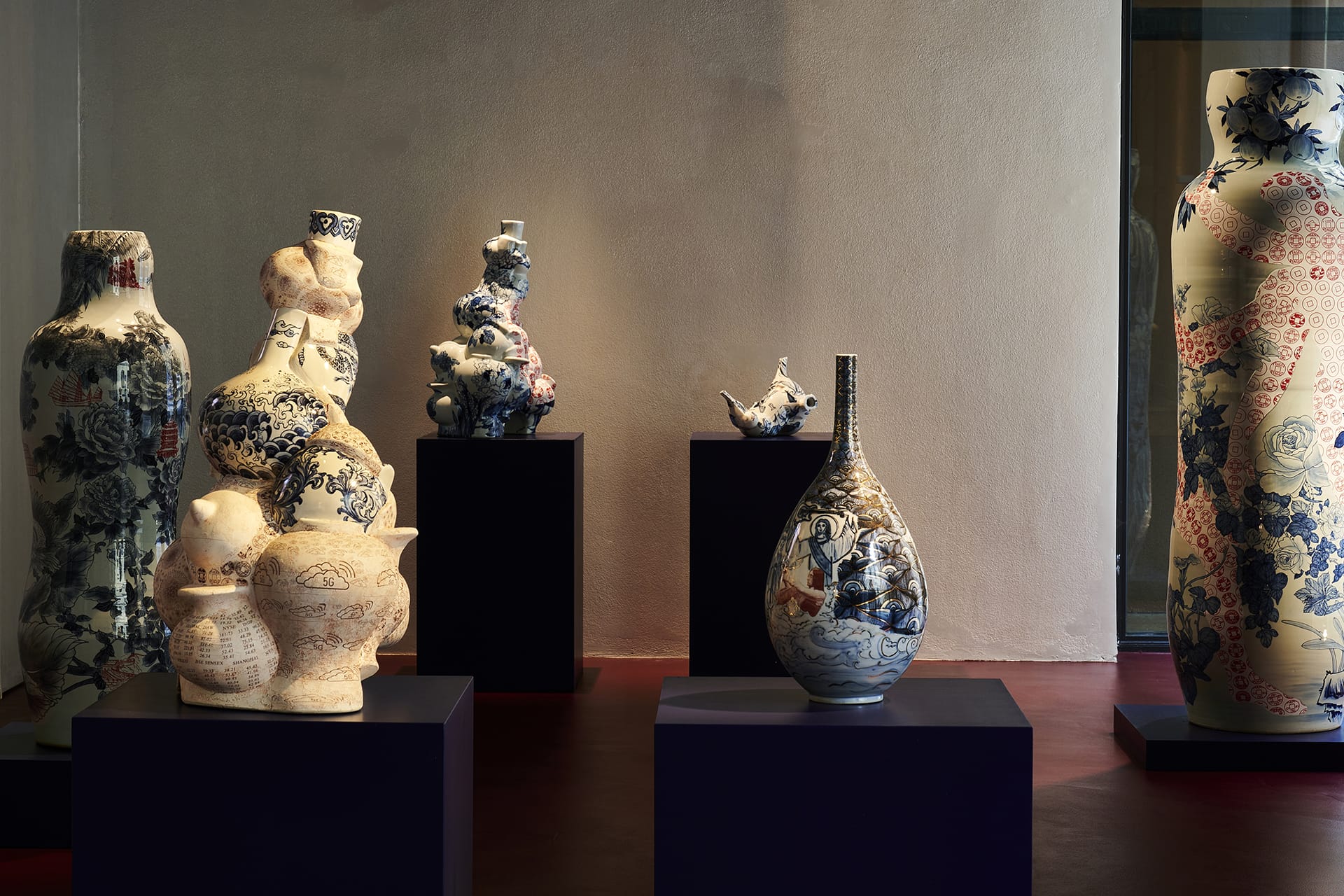 Diverse ceramic vases with intricate blue and red patterns displayed on dark pedestals in a dimly lit room.