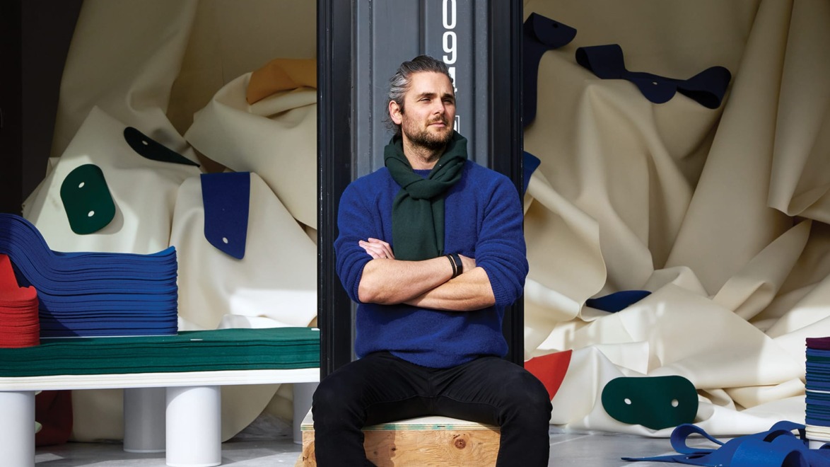 Person sitting on a wooden crate in a room filled with colorful fabric piles and abstract shapes in the background