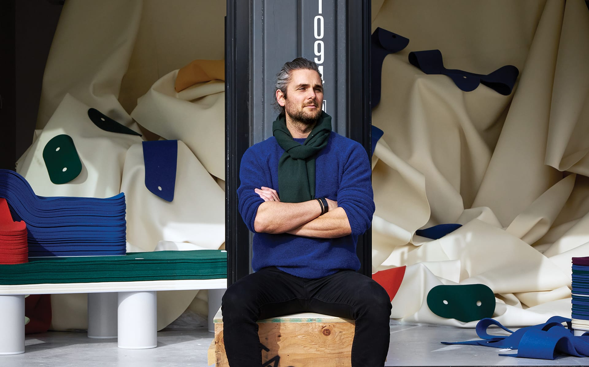 Person sitting on a wooden crate in a room filled with colorful fabric piles and abstract shapes in the background
