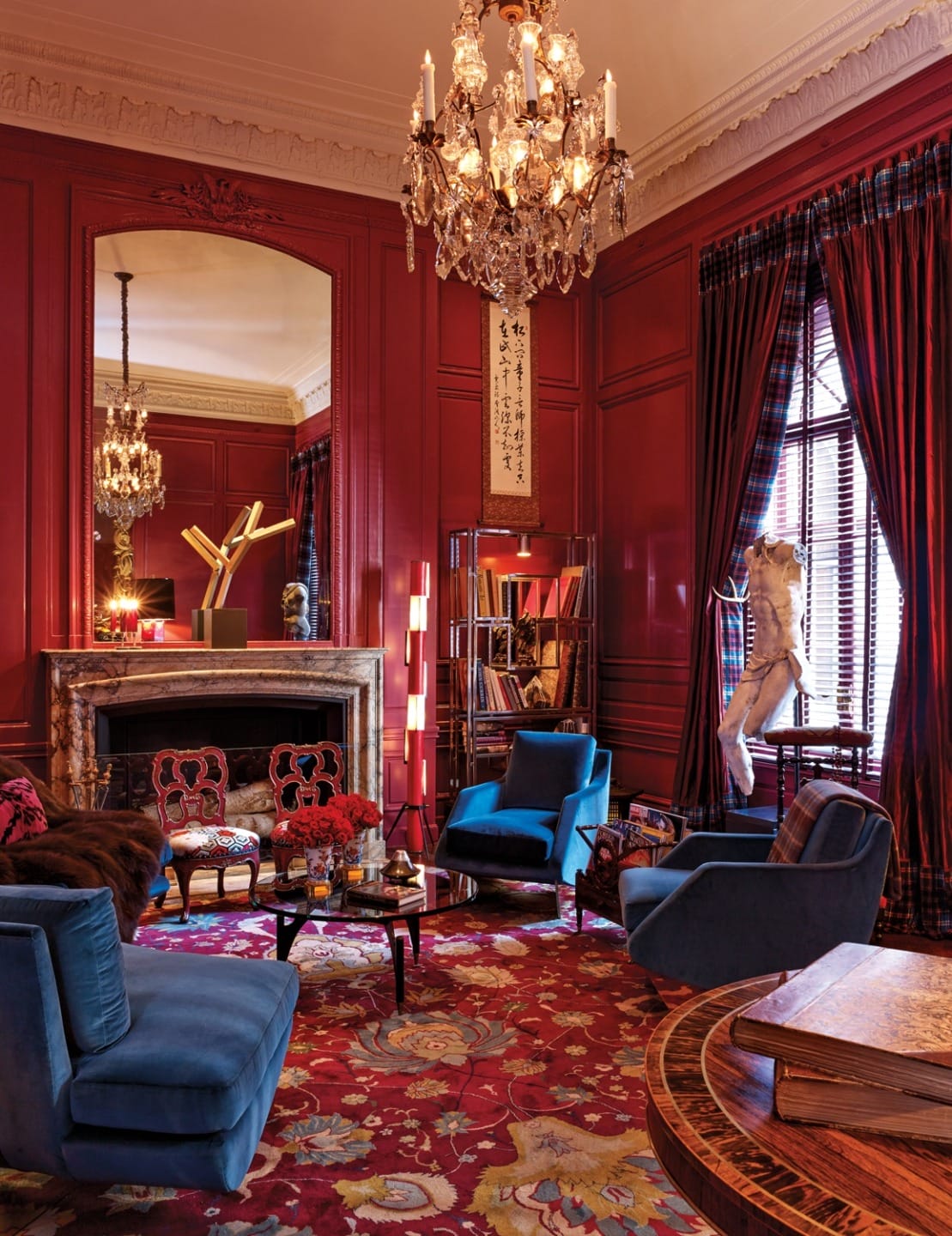 Luxurious living room with blue armchairs, red walls, chandelier, large mirror, and decorative sculpture on the mantel.