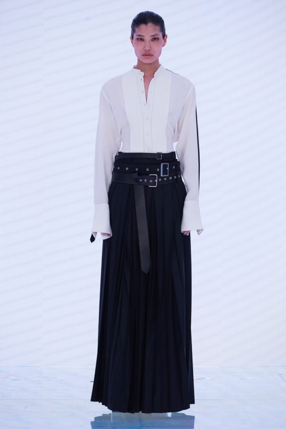 Model wearing a white blouse and long black skirt with a wide belt on a minimalist white background.