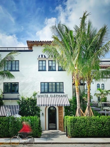 Tropical hotel exterior with palm trees, white facade, striped awnings, and a cyclist riding by on a sunny day.