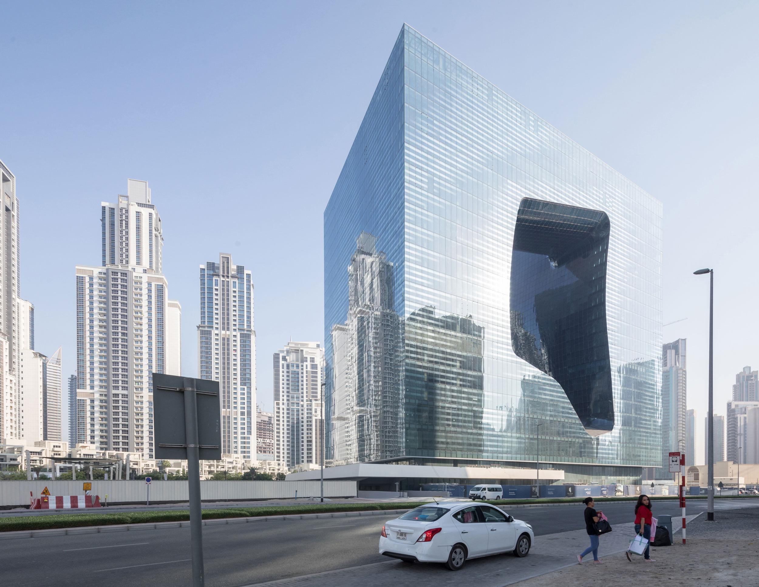 Modern glass building with a unique geometric cutout surrounded by skyscrapers and cars on a sunny day.
