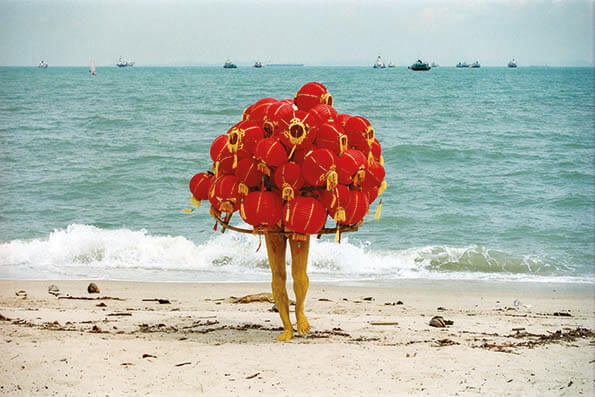Person standing on a beach covered in numerous red lanterns with ocean and boats in the background.