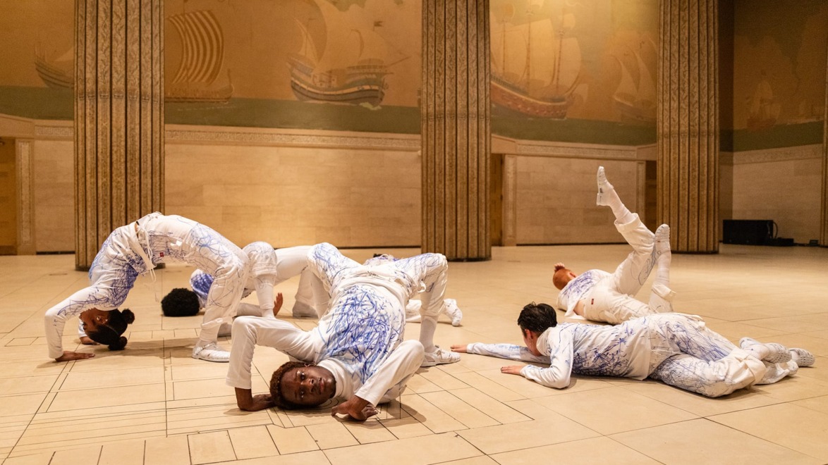 Dancers in artistic poses on a patterned floor with murals and tall columns in the background.