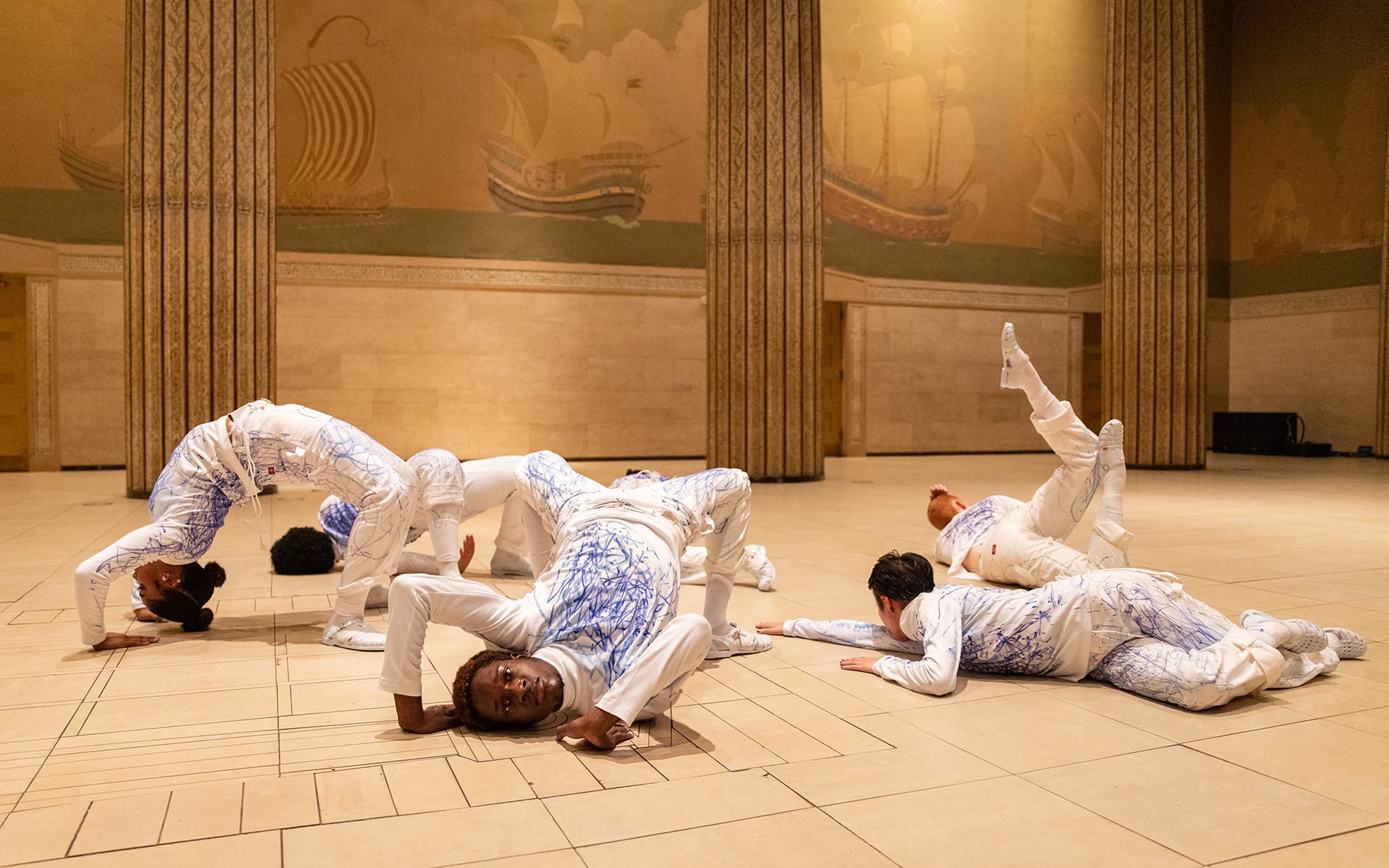 Dancers in artistic poses on a patterned floor with murals and tall columns in the background.
