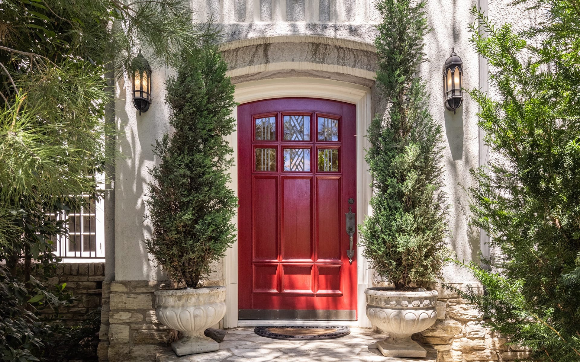 Red wooden front door framed by tall potted plants, set in a stone archway with decorative wall lanterns on each side.