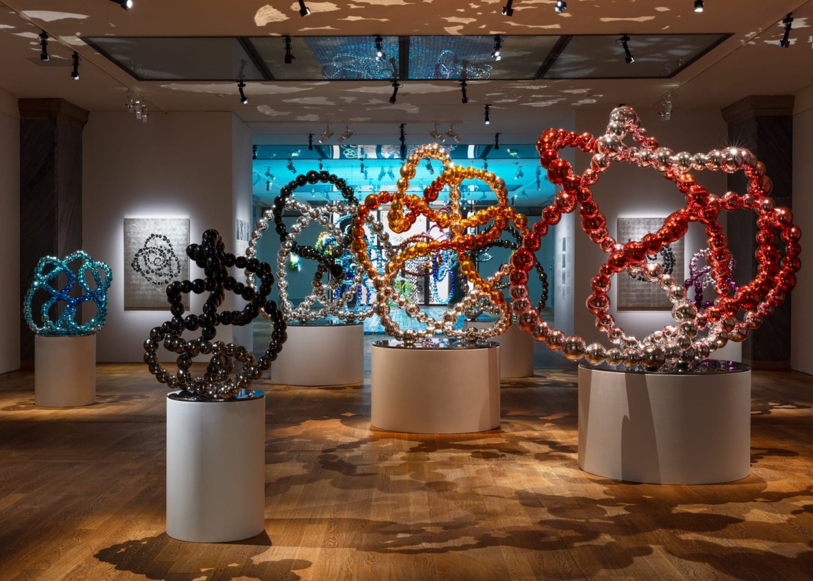 Colorful abstract sculptures displayed in a gallery with intricate patterns and lighting effects creating shadows on the walls.