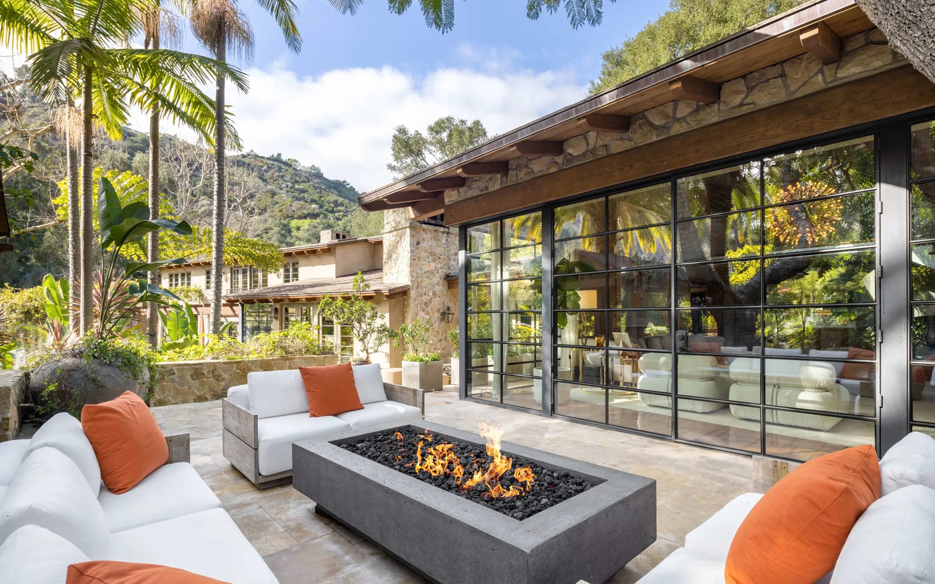 Modern patio with white sofas, orange cushions, and a fire pit; surrounded by lush greenery and mountains in the background.