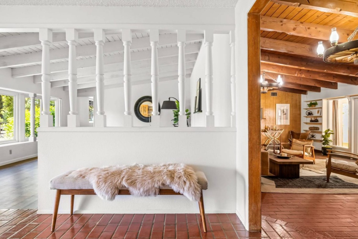 Interior view with a fur-covered bench on red tiles beside a room featuring wooden beams and large windows with greenery outside.