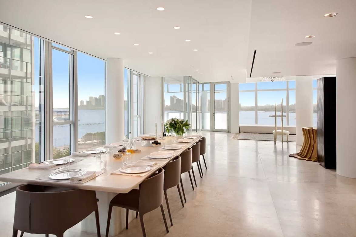Spacious dining area with a long table, modern chairs, and large windows offering a view of the city skyline and waterfront.