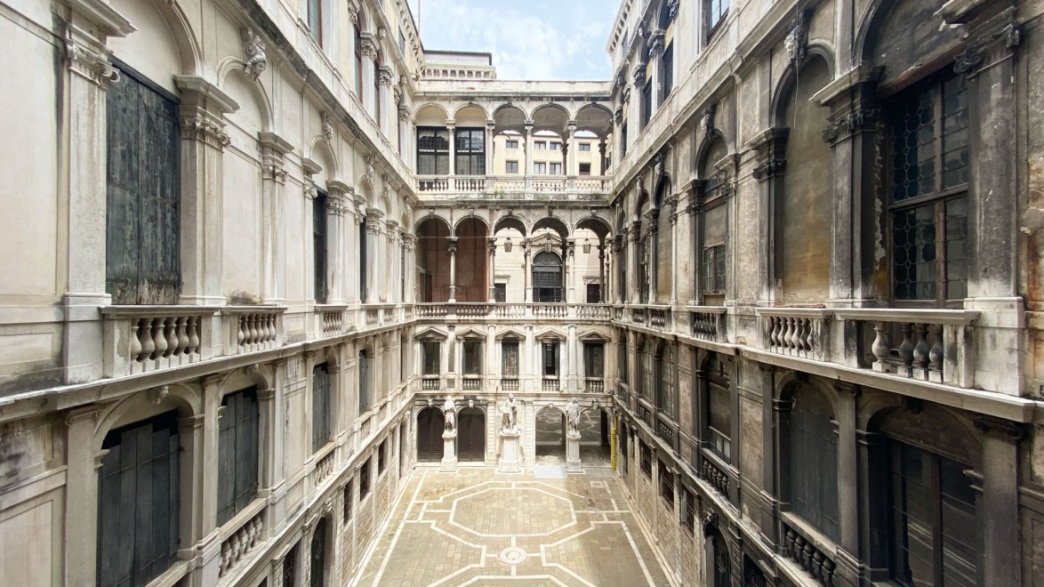 Interior view of a historic building's courtyard with balconies, arches, and a paved ground under a blue sky.