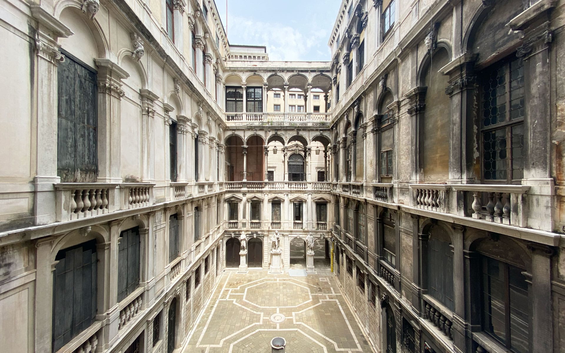 Interior view of a historic building's courtyard with balconies, arches, and a paved ground under a blue sky.