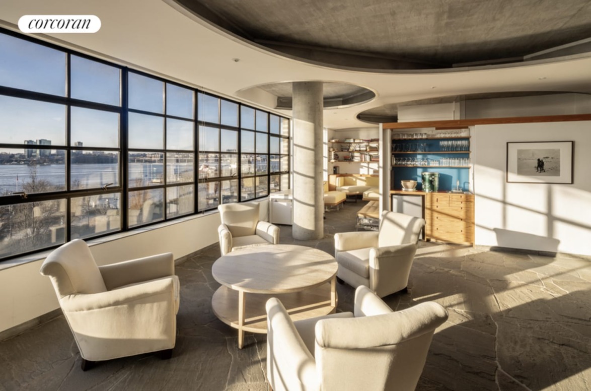 Spacious room with large windows, four armchairs around a round table, sunlight streaming in, view of waterfront outside