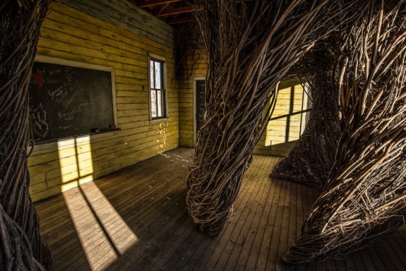 Abandoned room with twig structures, wooden walls, chalkboard with writings, and sunlight streaming through windows.