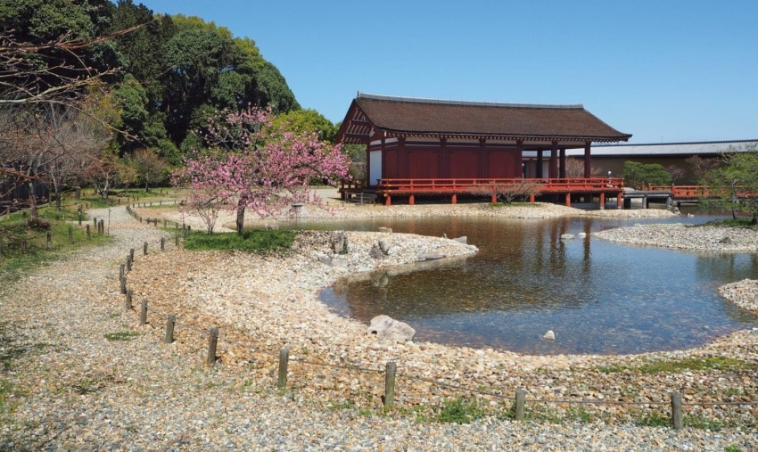 Japanese garden with a traditional wooden pavilion, cherry blossoms, pond, and stone pathway under a clear blue sky.
