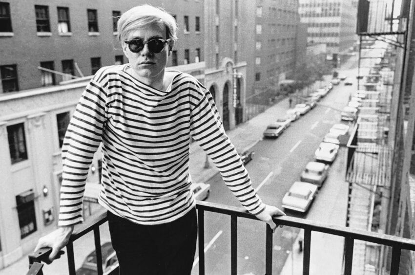 Person in striped shirt and sunglasses standing on a balcony overlooking a city street with cars and buildings.