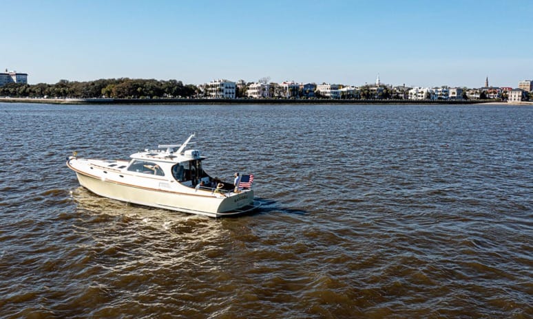 A white boat with an American flag on a river, city skyline in the background, under a clear blue sky.