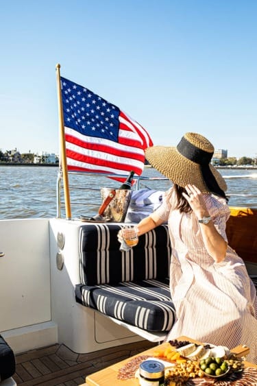 Person in a sun hat enjoying a drink on a boat with an American flag and snacks, against a sunny waterside backdrop.