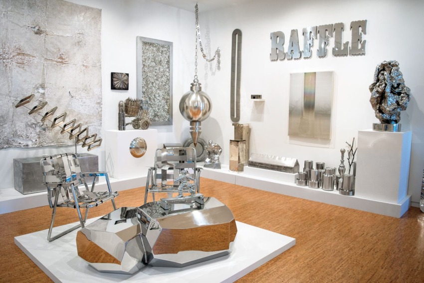 Contemporary art gallery with metallic sculptures and abstract installations against a white wall and wooden floor.