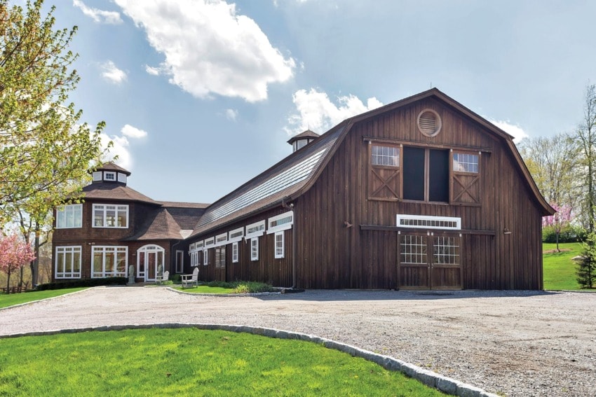 Large rustic barn with adjacent house, surrounded by trees and clear blue sky in the background.