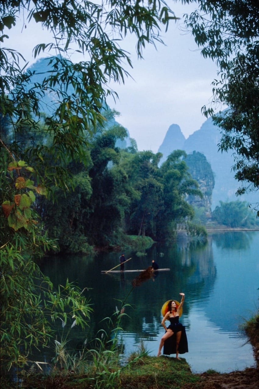 Person posing by a scenic river, surrounded by lush greenery and distant mountains, with two boats on the water.