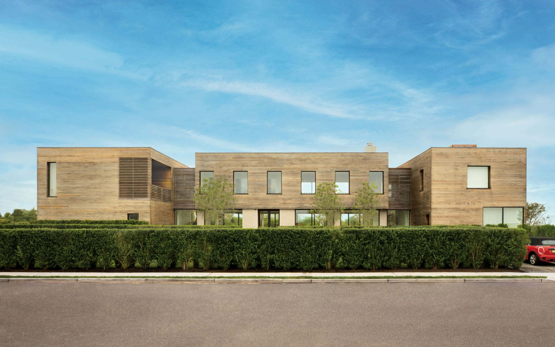 Modern wooden house with large windows, surrounded by greenery and a neatly trimmed hedge under a clear blue sky.