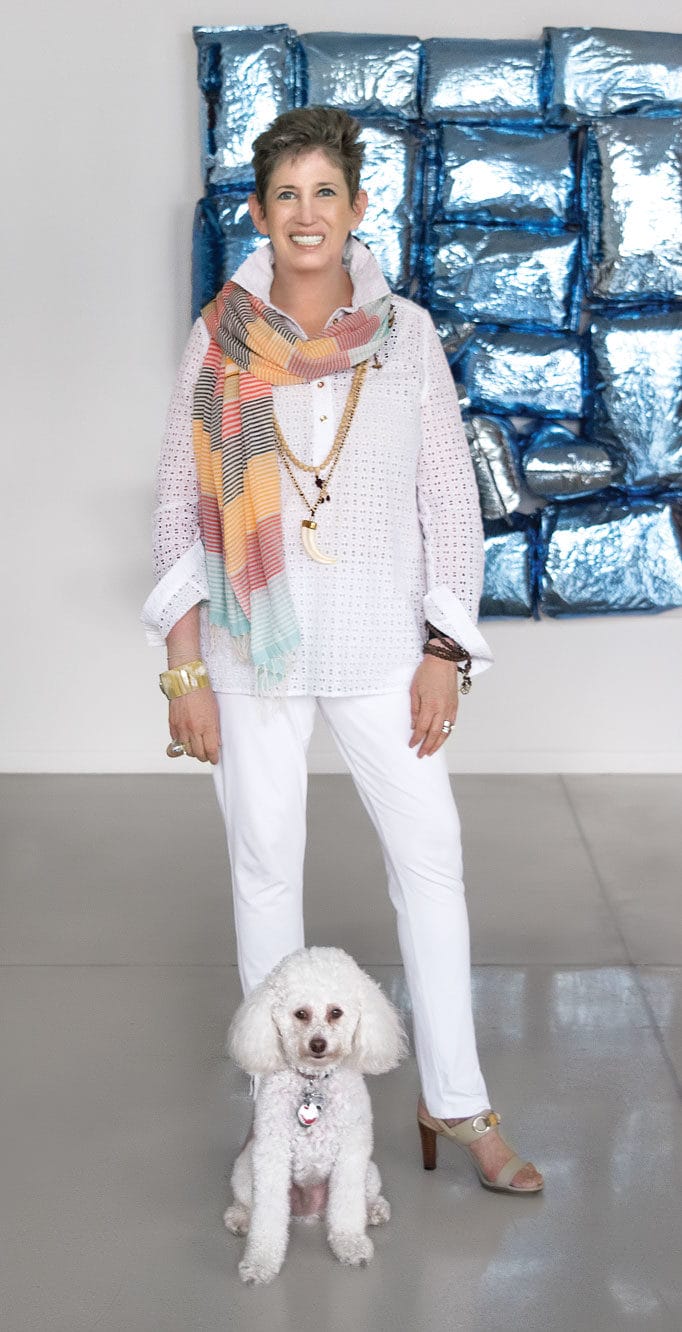Woman in white outfit with colorful scarf stands smiling next to small white poodle in front of blue abstract art installation