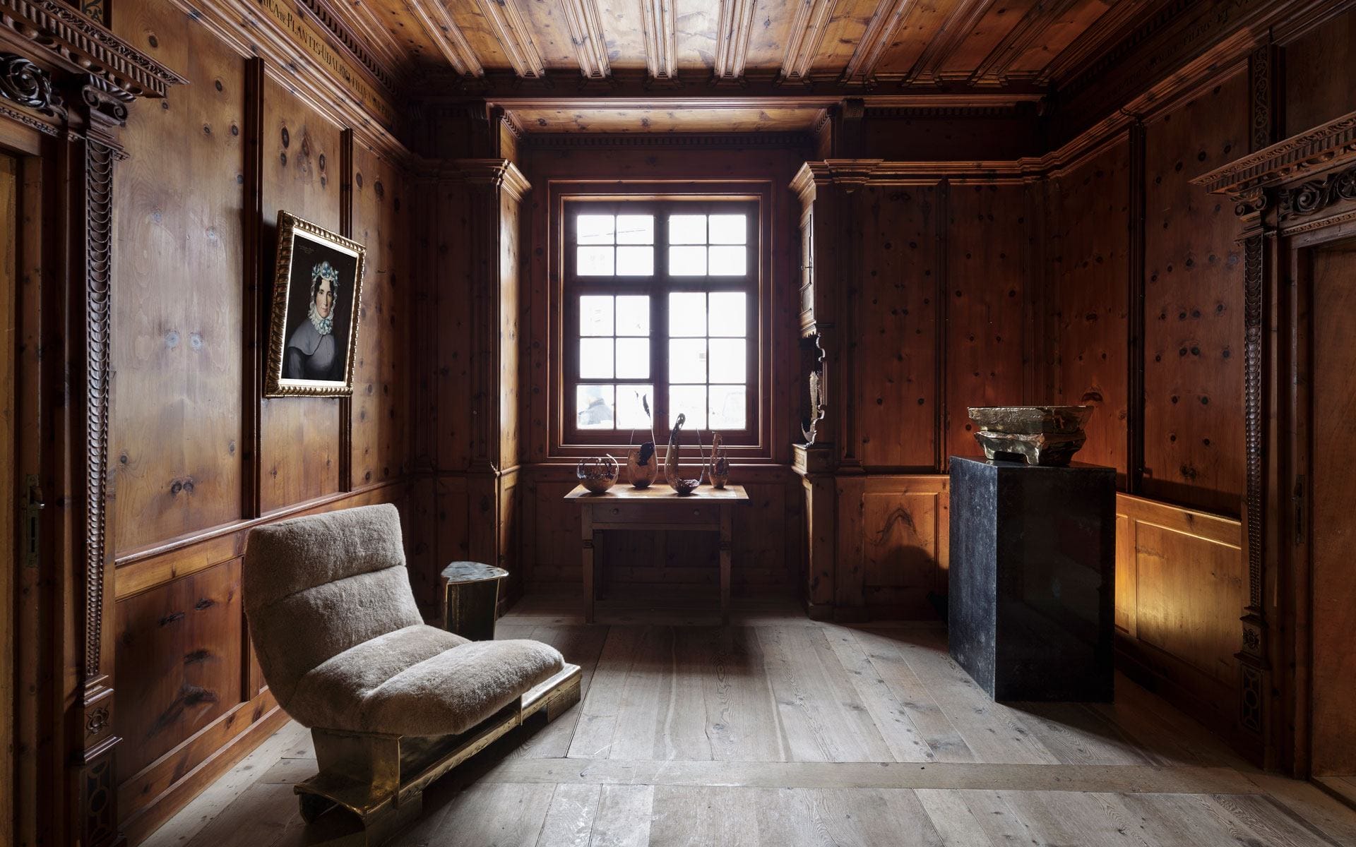 Wood-paneled room with a large window, an armchair, a pedestal display, and a painting on the wall.