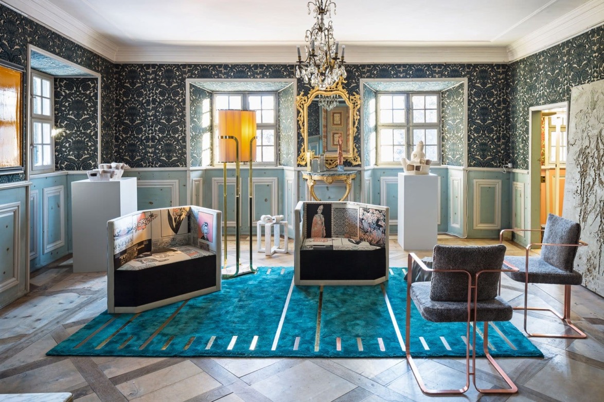 Elegant room with ornate chandelier, patterned walls, blue rug, modern sculptures, and contemporary chairs.